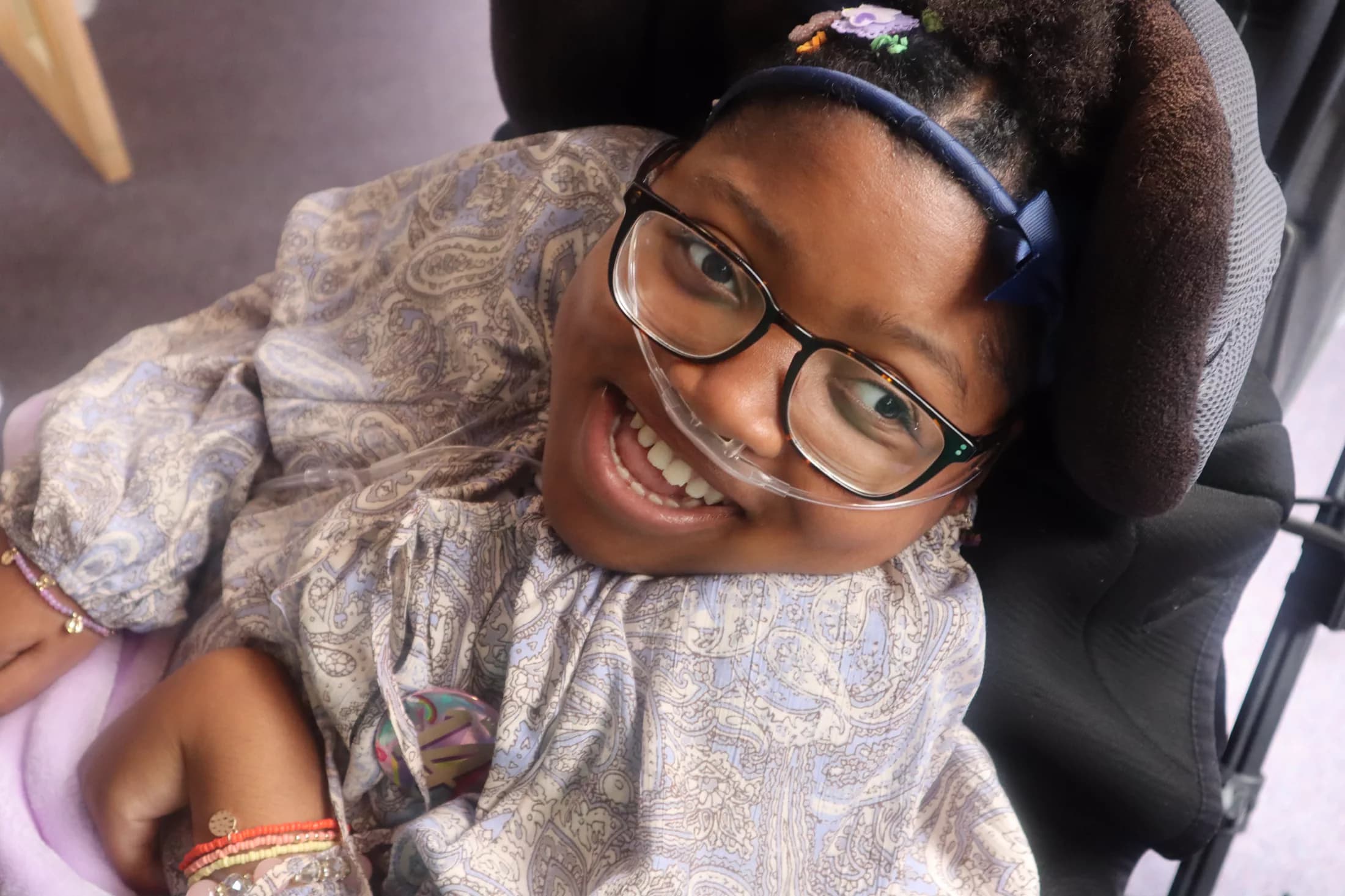 Abigail smiling brightly in her wheelchair, wearing glasses and a patterned headband.